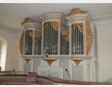 Historische Orgel in einem Kirchenraum, mit reich verzierten, goldenen Ornamenten und silberfarbenen Pfeifen, eingebettet in ein weiß-graues Gehäuse. Im Vordergrund ist eine Kirchenbank mit einer Tafelnummer und ein Mikrofon zu sehen.