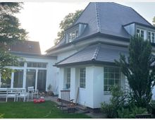 Weiße Einfamilienhaus mit dunklem Walmdach und Dachgauben, umgeben von einem Garten mit Bäumen, Rasen und Gartenmöbeln. Große Fenster und eine Glastür führen zum Außenbereich, wo ein Kinderfahrzeug und Stühle stehen.