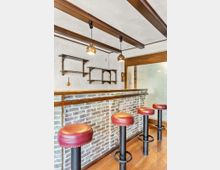 Innenraum einer kleinen Bar mit gemauerter Theke, Holzbalkendecke und zwei hängenden Kugelleuchten. Vor der Theke stehen mehrere hohe Barhocker mit roten Lederpolstern; an der weißen Wand sind offene Regale und rechts eine Milchglasschiebetür.