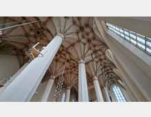 Blick nach oben in das Innere einer gotischen Kirche mit hohen weißen Pfeilern und einem reich gegliederten Kreuzrippengewölbe mit ornamentalen Schlusssteinen. Rechts fällt Licht durch große, hohe Fenster, darunter Spitzbogenfenster im Seitenschiff.