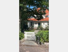 Ein zweistöckiges Haus mit weißer Fassade und rotem Ziegeldach, umgeben von einem gepflegten Garten mit Bäumen und Sträuchern. Im Vordergrund ist ein weißes Gartentor mit der Nummer „4A“ zu sehen, der Weg dahinter führt zum Eingang des Hauses.