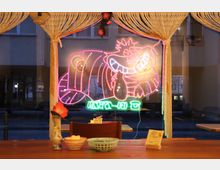 Ein Caféfenster mit einer neonbeleuchteten Illustration einer grinsenden Katze. Im Vordergrund sind ein Holztisch, kleine Schalen und eine Vase mit einer roten Blume zu sehen, eingerahmt von geflochtenem Dekor.