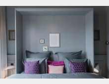 Graues Schlafzimmer mit einem Bett, das von einem eingerahmten Wandbereich umgeben ist. Das Bett ist mit grauer Bettwäsche, rosa und lila Kissen dekoriert; an der Wand hängen kleine gerahmte Bilder.