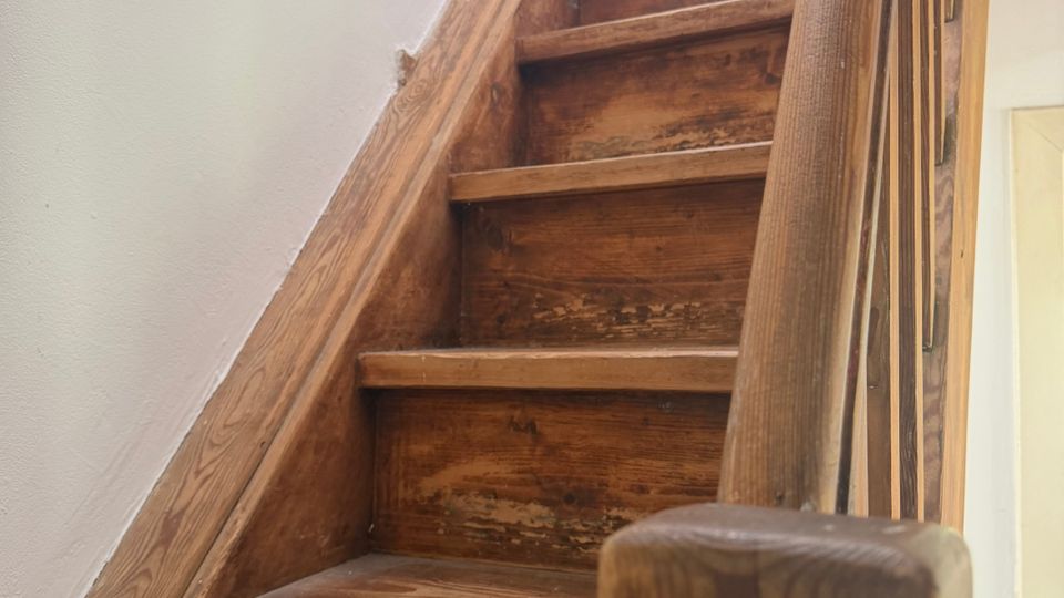 Schmale Holztreppe in einem Innenraum, mit abgenutzten Stufen und einem Holzgeländer an der rechten Seite. Weiß gestrichene Wände und ein helles Fenster bzw. Lichtöffnung oben am Treppenlauf.