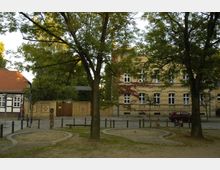 Straßenszene mit einem historischen Backsteingebäude und einem Fachwerkhaus im Hintergrund, umgeben von Bäumen. Im Vordergrund befinden sich ein kleiner Sandspielbereich und gepflasterte Wege.