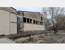 Verwittertes Industrie- oder Lagergebäude mit Wellblechdach, abblätternder Fassade, großen Holztoren und einer Reihe schmaler Fenster über einer erhöhten Laderampe. Davor liegt eine breite Betonfläche mit etwas Bauschutt; im Hintergrund stehen kahle Bäume.