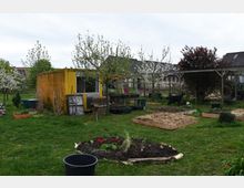 Ein Gemeinschaftsgarten mit Hochbeeten, blühenden Obstbäumen und einem gelben Containergebäude im Hintergrund. Im Vordergrund sind ein bepflanztes Beet und eine schwarze Plastiktonne auf einer Grünfläche zu sehen, umgeben von weiteren Gartenstrukturen.