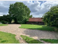 Kopfsteinpflasterweg in einer ländlichen Umgebung, umgeben von gepflegten Rasenflächen und üppigen Bäumen. Im Hintergrund steht ein rustikales Gebäude mit roten Ziegeldach, flankiert von dichten Hecken und einem Tor.