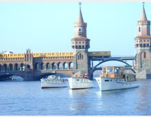 Die Aufnahme zeigt die Oberbaumbrücke in Berlin, eine historische Brücke mit zwei Türmen aus Backstein und Bögen, über der eine gelbe U-Bahn fährt. Im Vordergrund schwimmen mehrere weiße Boote auf dem Wasser der Spree.