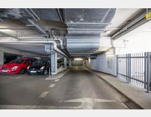 Ein Tiefgaragenbereich mit geparkten Autos auf der linken Seite, rechteckigen Lüftungskanälen und Rohren an der Decke. Rechts befinden sich ein Metallgitterzaun und eine Fahrbahn mit Pfeilmarkierungen, die auf eine Ausfahrt hinweisen.