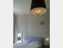 Ein Schlafzimmer mit einem gepolsterten Doppelbett, weißer Bettwäsche und einer hellgrauen Wand. Im Raum befinden sich ein großer schwarzer Kronleuchter mit Kerzenlampen, ein kleiner Beistelltisch mit einer Lampe und ein bodenstehender Spiegel neben einem Fenster mit Vorhängen.