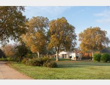 Neoklassizistisches Gebäude mit Säulenfassade, umgeben von großen Bäumen mit herbstlichem Laub, in einer weitläufigen Parklandschaft mit Rasenflächen und einem Kiesweg im Vordergrund.