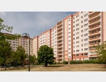 Mehrstöckiges Wohngebäude im Stil eines Plattenbaus mit mehreren Balkonen, umgeben von einer Grünfläche mit Bäumen, Bänken und einem geschwungenen Straßenlaternenmast im Vordergrund. Der Himmel ist leicht bewölkt.