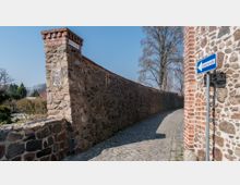 Teil einer historischen Stadtmauer aus unregelmäßigen Natursteinen mit Ziegelabschlüssen, daneben eine gepflasterte Gasse. Ein Straßenschild weist auf eine Einbahnstraße hin, Bäume und Büsche im Hintergrund.