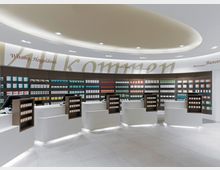 Innenraum einer modernen Apotheke mit einer geschwungenen Decke und heller Beleuchtung. Der Raum zeigt mehrere weiße Verkaufstheken mit integriertem Regalsystem, dahinter befinden sich hohe Regale mit Medikamentenpackungen.