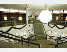 Innenansicht eines öffentlichen Gebäudes mit Treppenhaus und dekorierten Geländern, geschmückt mit Weihnachtsschmuck und Girlanden. Im oberen Bereich befinden sich großflächige Fenster, ein geschmückter Weihnachtsbaum und Kunstwerke an den Wänden; eine große Leuchte hängt zentral im Raum.