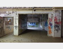 Ein verlassener, heruntergekommener Raum mit beschädigten Wänden und Decken, gefliest im unteren Bereich, mit Graffiti bedeckt. Der Boden ist mit Blättern übersät, und Rohre verlaufen unter der Decke.