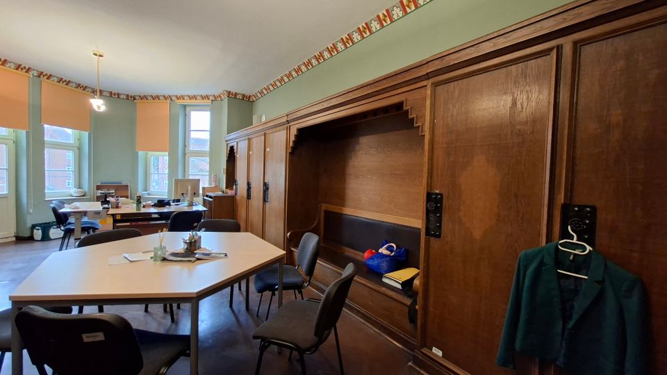 Innenraum eines Arbeits- oder Besprechungszimmers mit mehreren Tischen und Stühlen sowie hohen Fenstern mit heruntergelassenen Rollos. Entlang der rechten Wand steht eine große, dunkle Holzvertäfelung mit integrierter Sitznische und Schrankelementen; die Wände sind hellgrün mit dekorativem Bordürenfries.