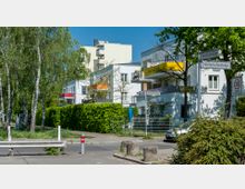 Wohngebiet mit modernen Gebäuden, die helle Fassaden und bunte Balkone aufweisen. Im Vordergrund sind eine Baumreihe, grüne Hecken, Straßenschilder und eine Verkehrsberuhigungszone mit einer Geschwindigkeitsbegrenzung von 30 km/h zu sehen.