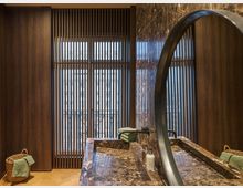 Modernes Badezimmer mit dunklen Holzpaneelen, bodentiefen Fenstern mit vertikalen Lamellen, einer Waschtischplatte aus poliertem braunem Marmor und einem großen, runden Spiegel. Ein geflochtener Korb mit Handtuch und weitere grüne Handtücher ergänzen die Einrichtung.