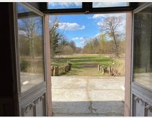 Blick durch eine geöffnete, verglaste Doppeltür auf eine breite Betontreppe und einen flachen, grasbewachsenen Weg, der in eine Wiese führt. Im Hintergrund stehen kahle und teilweise belaubte Bäume unter blauem Himmel mit einigen Wolken.