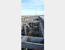 Ansicht eines industriellen Stahlgerüsts mit rostigen Trägern, aufgenommen von einer erhöhten Position. Im Hintergrund ist eine flache Landschaft mit blauem Himmel und einigen dünnen Antennenmasten zu sehen.