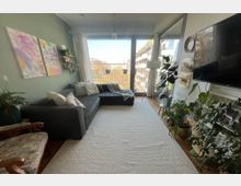 Wohnzimmer mit großer Fensterfront, die auf einen Balkon mit Stadtblick führt. Der Raum ist eingerichtet mit einem dunkelgrauen Ecksofa, Teppichboden, mehreren Zimmerpflanzen und dekorativen Elementen an der Wand.