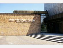 Außenansicht des Zeiss-Großplanetariums in Berlin, mit einer Fassade aus gelblich-braunen Klinkersteinen, breiten Fensterfronten und einem Teil der silbernen Kuppelstruktur im Hintergrund. Rechts führen halbrunde Stufen zu einem Eingang.