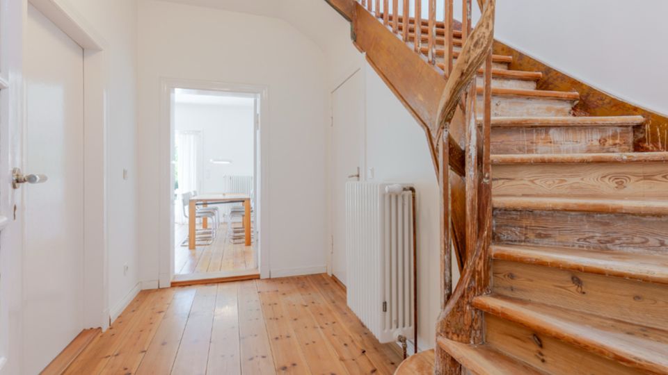 Heller Flur in einem Wohnhaus mit Holzdielenboden und einer geschwungenen Holztreppe mit Geländer an der rechten Seite. Durch eine Türöffnung ist ein angrenzender Raum mit Tisch und Stühlen sichtbar; an der Wand steht ein weißer Heizkörper.