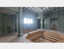 Ein leerer, verlassener Raum mit einem eingelassenen Becken in der Mitte, umgeben von marmorglänzenden, hellgrünen Wandfliesen. Eine große Fensteröffnung mit Gitter und ein Heizkörper befinden sich an der Wand, während die Decke mit sichtbaren Betonbalken gestaltet ist.