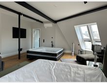 Modernes Dachgeschoss-Schlafzimmer mit sichtbaren schwarzen Stahlträgern und schrägen Wänden, in der Mitte mit einer großen Whirlpool-Badewanne auf dunklem Sockel. Rechts sorgen große Dachflächenfenster für Tageslicht; Holzboden, TV an der Wand und wenige Möbel ergänzen den Raum.