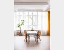 Heller Raum mit großen Sprossenfenstern, weißen Vorhängen und gelbem Vorhang an der rechten Seite. In der Mitte steht ein schwarzer Tisch mit sechs Holzstühlen, links eine Zimmerpflanze in einem Topf auf hellem Holzboden.