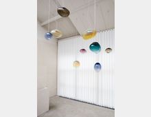 Minimalistischer Innenraum mit hellen Wänden, einem glatten Betonboden und einer hohen Fensterfront, die mit weißen Vorhängen bedeckt ist. Von der Decke hängen mehrere bunte, kugelförmige Glaslichter an dünnen Kabeln.