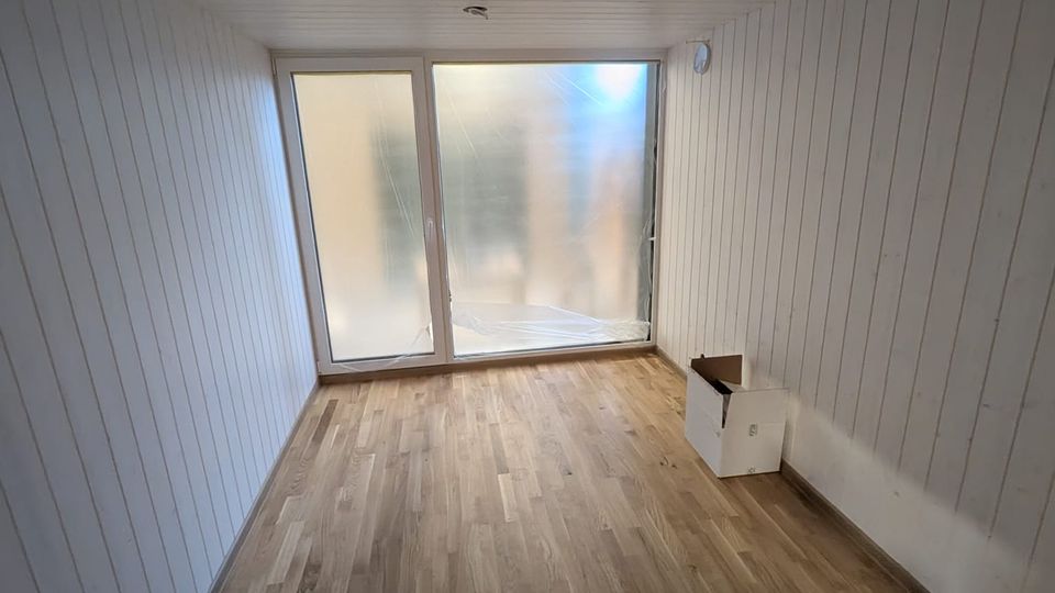 Schmaler, leerer Innenraum mit weiß vertäfelten Wänden und Decke sowie Holzfußboden. Am Ende befindet sich eine große, milchig verglaste Schiebetür bzw. Fensterfront, die Tageslicht in den Raum lässt.