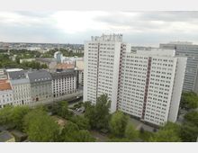 Blick auf eine städtische Umgebung mit einem großen weißen Hochhaus mit roten Streifen und vielen Fenstern im Vordergrund. Umgeben wird das Gebäude von weiteren Wohnhäusern, kleineren Gebäuden im klassischen Baustil und einer parkähnlichen Grünfläche mit Bäumen und einzelnen Wegen.