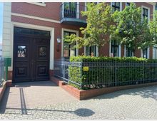 Rotes Backsteingebäude mit dunkelbrauner Holztür und verzierten Fenstergittern. Vor dem Gebäude befinden sich ein schmiedeeiserner Zaun, eine niedrige Hecke und gepflasterter Gehweg mit einem Hinweisschild „Ausfahrt freihalten“.