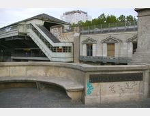 Steinbrücke mit der Inschrift „Hallesche-Tor-Brücke“ im Vordergrund, dahinter ein moderner U-Bahn-Eingang mit metallischen und gläsernen Elementen sowie ein historisch anmutendes Gebäude aus Stein mit rechteckigen Fenstern. Im Hintergrund sind Bäume und ein teilweise verdecktes Gebäude mit Gerüst zu sehen.