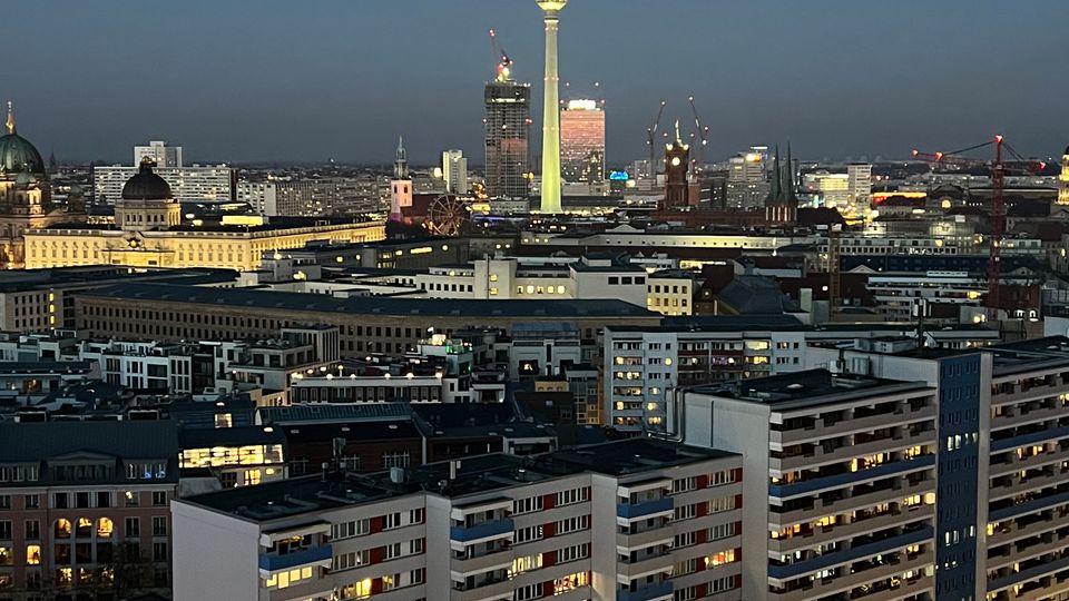 Abendansicht einer städtischen Skyline in Berlin mit dem beleuchteten Berliner Fernsehturm im Zentrum. Im Vordergrund befinden sich mehrstöckige Wohngebäude, Straßenlaternen und ein Einkaufsbereich, während der Himmel in der Dämmerung dunkelblau ist.