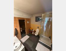 Modernes Badezimmer mit dunklen Bodenfliesen, Glasduschkabine und Toilette; links befindet sich eine holzverkleidete Sauna mit Glastür. Ein Waschbeckenunterschrank, ein kleines Wandfenster und ein Ablageregal mit Handtüchern ergänzen den Raum.