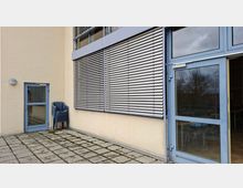 Außenbereich an einem modernen Gebäude mit hell verputzter Fassade, großen Fenstern mit horizontalen Außenjalousien und einer blau gerahmten Glastür. Der Boden ist mit quadratischen Betonplatten gepflastert; neben einer kleineren Nebentür stehen gestapelte Kunststoffstühle.