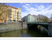 Mehrstöckige Wohngebäude in Pastellfarben stehen entlang eines Kanals mit einer steinernen Ufermauer. Eine verzierte grüne Metallbrücke führt über das Wasser, umgeben von kahlen Bäumen und einem teils bewölkten Himmel.