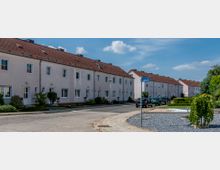 Reihenhaussiedlung mit weiß verputzten Fassaden und roten Ziegeldächern in einer ruhigen Wohnstraße. Im Vordergrund eine kleine bepflanzte Grünfläche und ein Straßenschild mit der Aufschrift „Grünstraße“, im Hintergrund geparkte Autos und blauer Himmel mit vereinzelten Wolken.