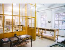 Heller Werkstattraum mit gelben Metallrahmen und Glaswänden, mehreren Holztischen mit Hockern und unterschiedlichen Arbeitsmaterialien. Große Fenster lassen Tageslicht hinein, und im Vordergrund steht ein Druckgerät auf einem Metalltisch.