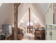 Dachgeschosszimmer mit schrägen weißen Wänden, einem zentralen Holzbalken, zwei Einzelbetten, Korbmöbeln und Stehlampen. Im Hintergrund ein halbkreisförmiges Fenster und ein kleiner Schreibtisch mit einem Korb darunter.