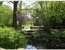 Kleiner Park mit einer Holzbrücke über einem Bach, umgeben von üppigen Bäumen und grünen Sträuchern. Im Hintergrund steht ein Gebäude mit einer Treppe und steinernen Elementen, eingerahmt von weiteren Pflanzen und einem Gartenbereich.