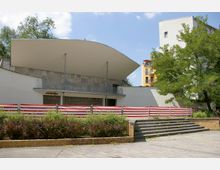 Außenansicht eines modernen Gebäudes mit geschwungenem Dach, Betonwänden und offener Treppe, umgeben von Bäumen und Sträuchern. Im Vordergrund ist ein rot-weiß gestreifter Zaun sichtbar, dahinter befindet sich ein gelber Wohnblock.