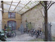 Ein gepflasterter Innenhof mit einem Baum, mehreren abgestellten Fahrrädern und einer Überdachung aus Glas und Metall. Im Hintergrund eine Backsteinfassade mit roter Tür sowie einem Oberlicht, daneben ein Zaun mit einem abstrakten Metallschild.