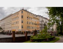 Straßenszene mit mehrstöckigen Wohngebäuden im Altbaustil, darunter ein markantes gelbes Eckhaus mit Geschäften im Erdgeschoss. Im Vordergrund ist ein kleiner, gepflegter Garten mit Beeten, Bäumen und einem Ziegelzaun zu sehen.