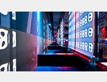 Langer schmaler Innenraum einer modernen Bar oder eines Clubs mit dunklen Sitzbereichen, beleuchtet von bunten Neonlichtern und leuchtenden Tafeln mit Zahlen- und Symbolmustern auf der rechten Seite. Der Raum zeigt eine urbane, industrielle Atmosphäre mit offenen Deckenstrukturen.