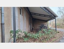 Überdachter Anbau an einem verputzten Gebäude mit Metallstützen und Wellblechdach; darunter steht eine lange, dunkle Sitzbank. Im Vordergrund wuchert Gestrüpp und Laub, daneben verläuft ein schmaler Weg am Rand eines baumreichen Grundstücks.